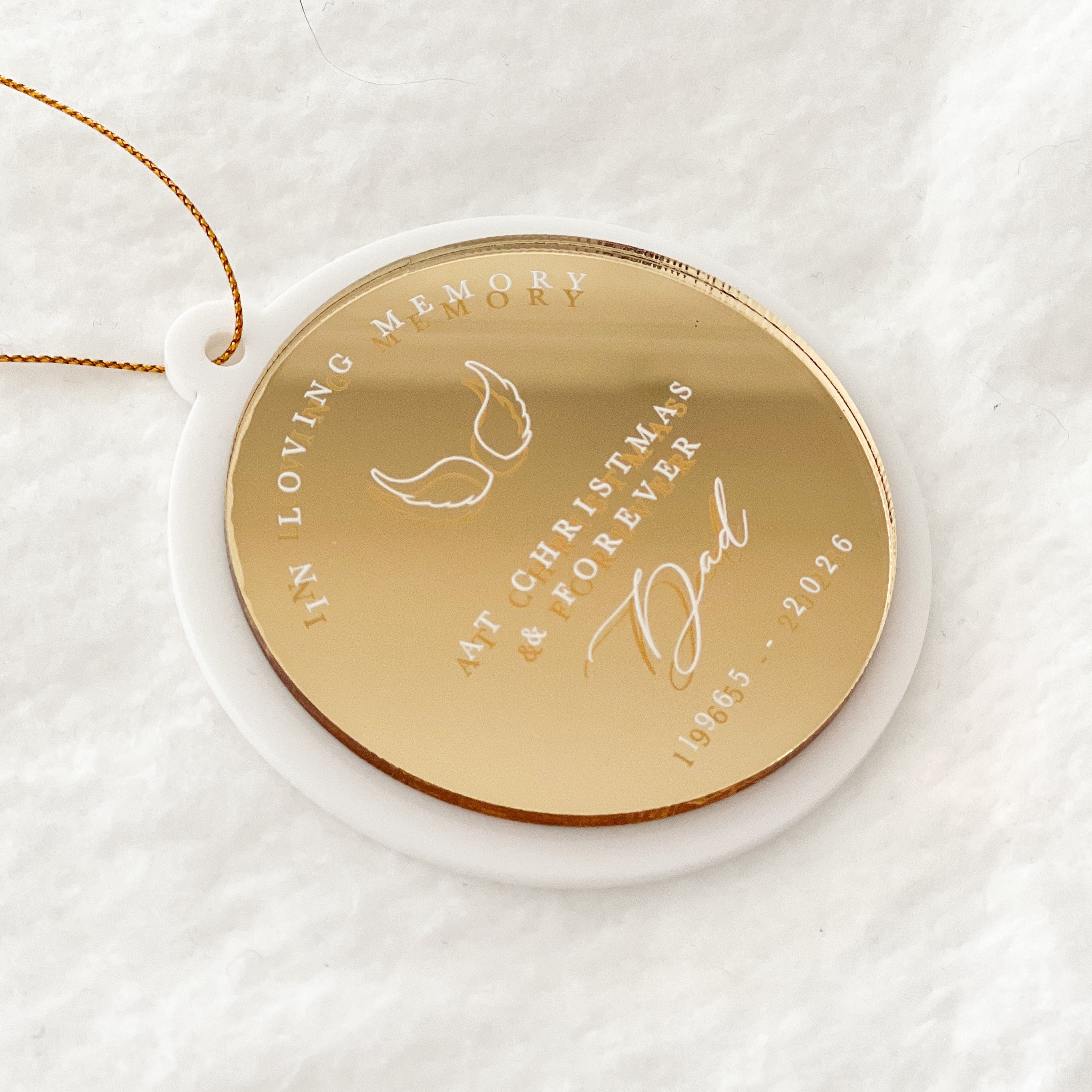 A round gold-toned memorial Christmas ornament with engraved text and a hanging loop, resting on a white circular base against a soft white textured background.