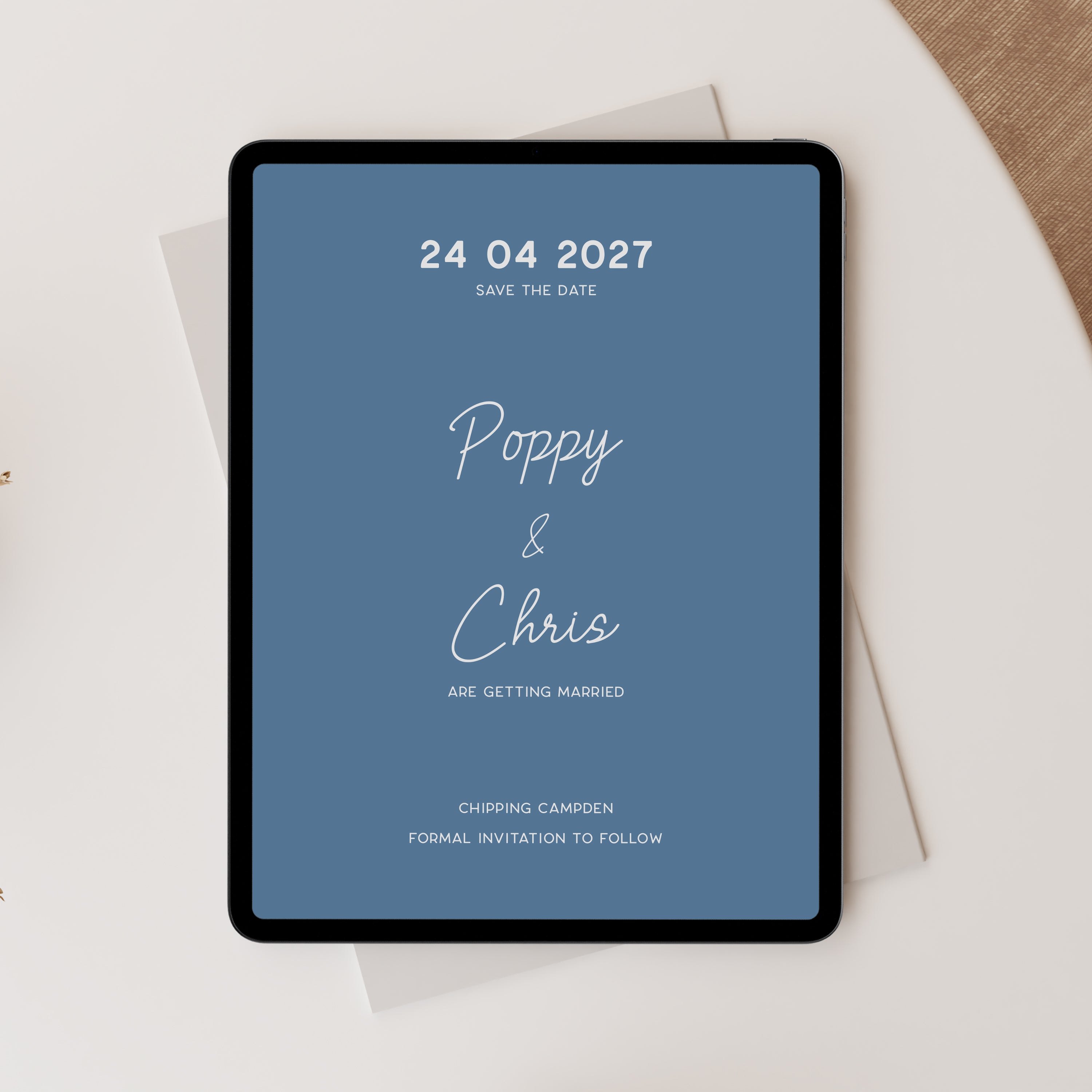 A tablet displaying a blue wedding save-the-date with white text, styled on top of white envelopes on a light beige surface.