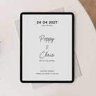 A black-framed digital save-the-date displayed on a light beige surface, with a white envelope behind it and a textured tan fabric corner in the upper right.