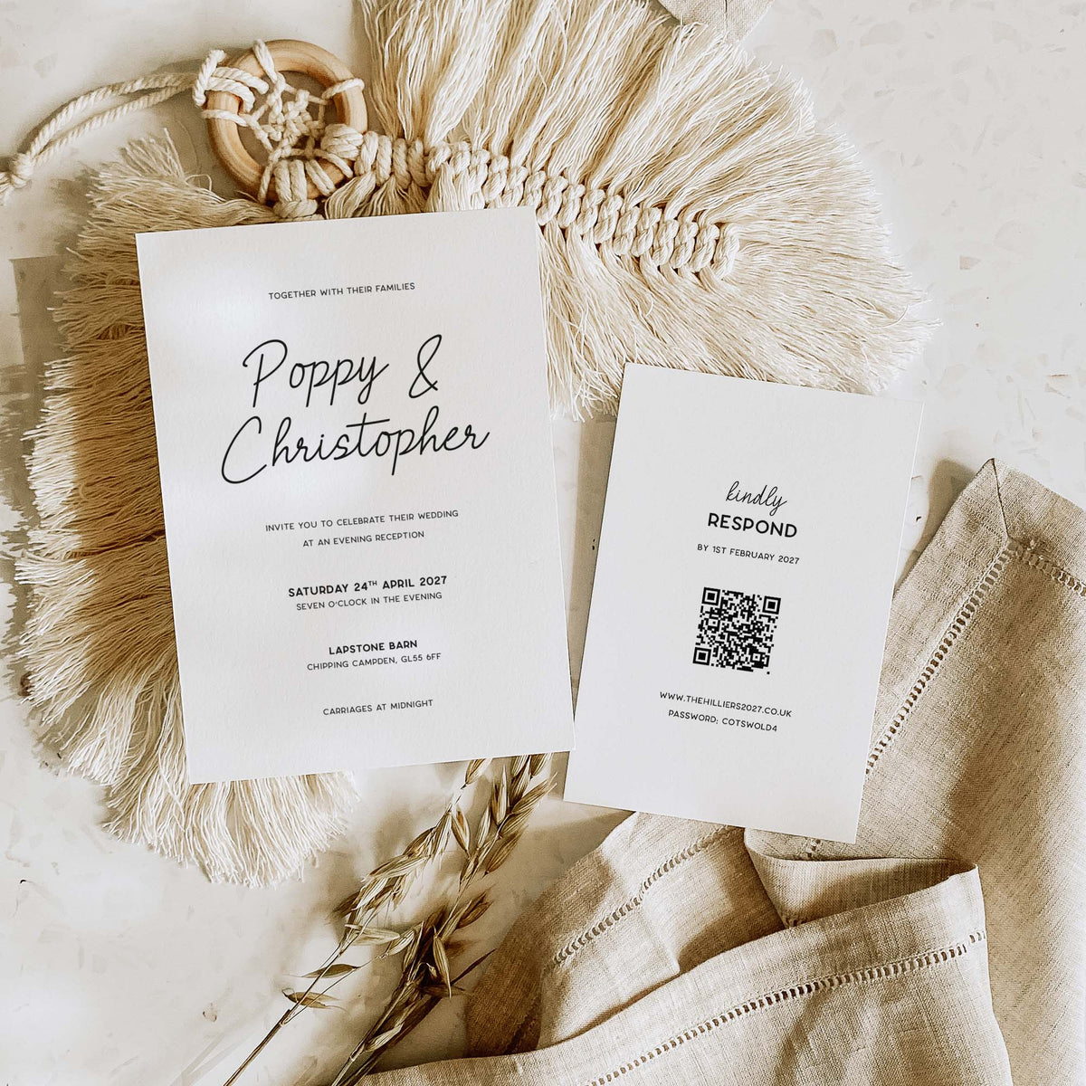 A styled flat-lay of wedding stationery on a neutral fabric surface. Two white cards are shown: a main invitation reading 'Poppy & Christopher' with wedding details, and a separate 'happy response' card with a QR code.