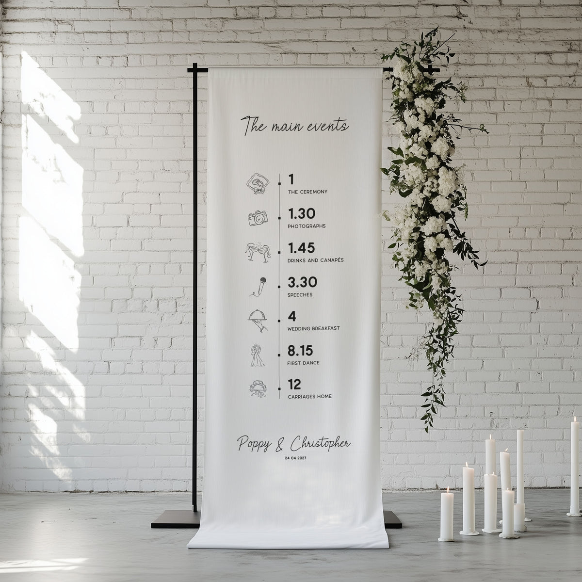A fabric wedding itinerary sign hanging from a black stand decorated with white florals and candles, in front of a white brick wall