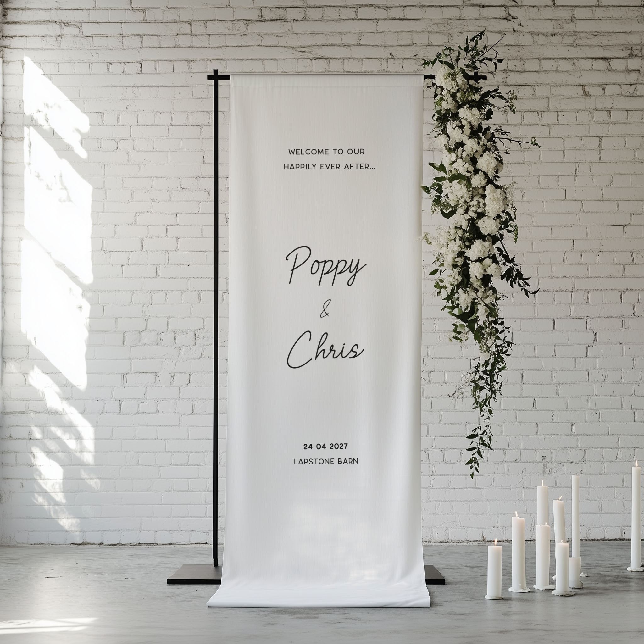 A vertical white fabric wedding backdrop banner on a black metal stand, displayed in a bright white brick room with floral greenery and white candles.