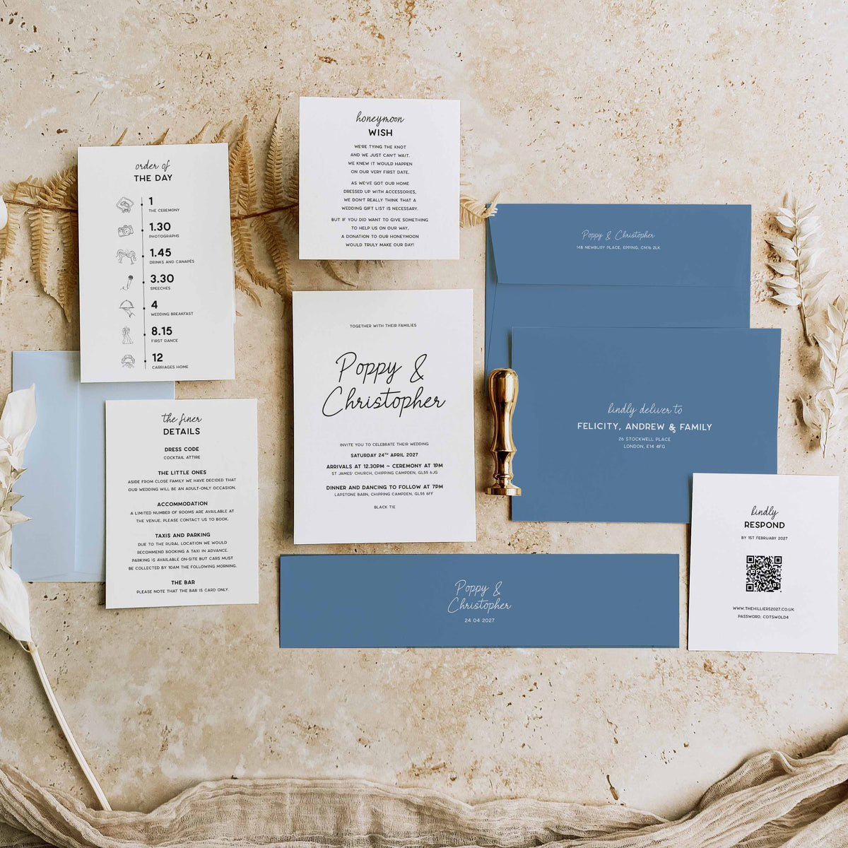 A coordinated wedding invitation suite arranged on a textured beige surface with dried botanicals. The set includes white cards with black and gray typography, muted blue envelopes, a blue card, a small blue card, a gold pen, and a QR code card.