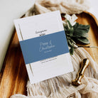 A stack of white stationery cards with a dusty blue band across the center, styled on a wooden tray with beige fabric, green leaves, and a gold wax seal stamp. The top card reads 'honeyman WISH' with names and date on the lower card.