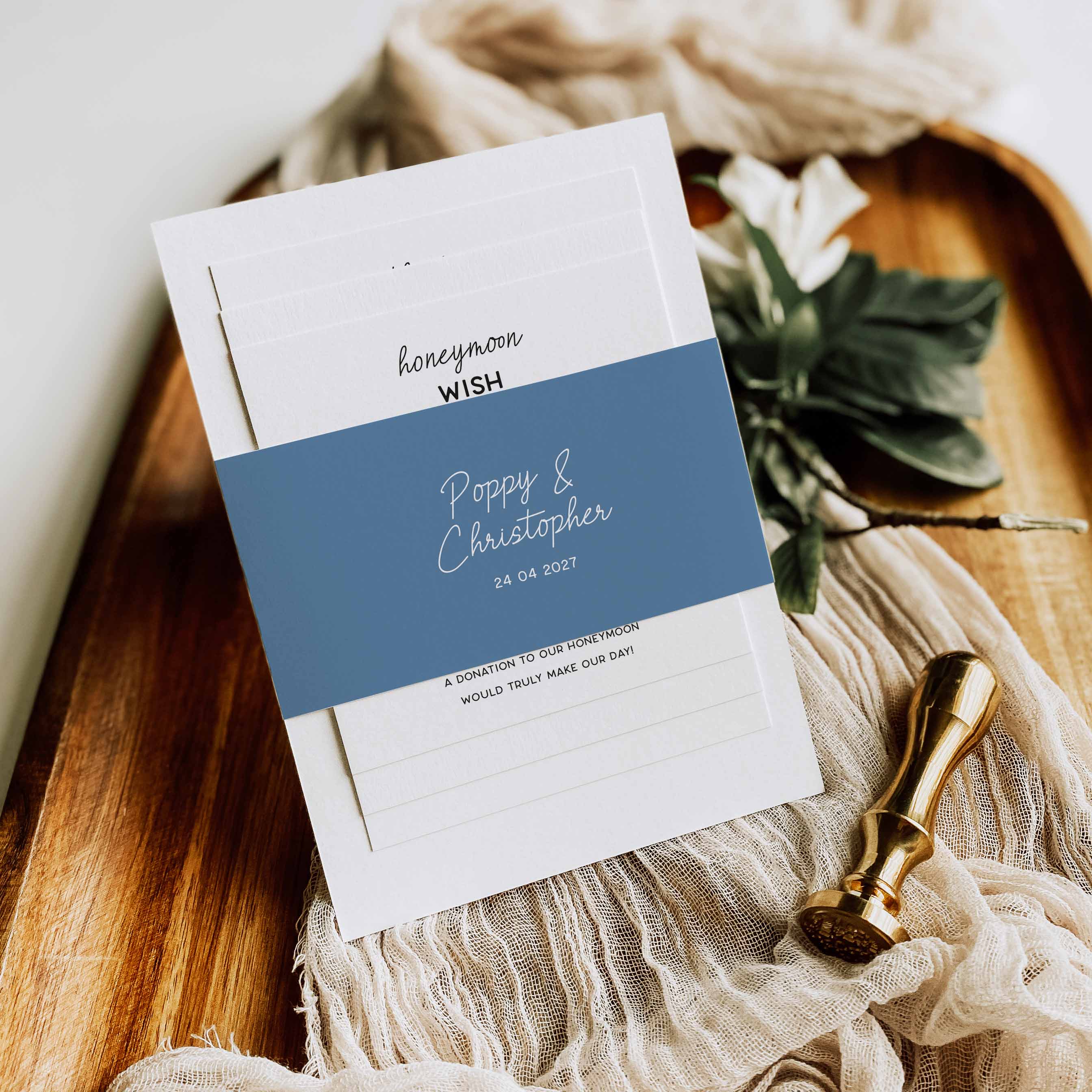 A stack of white stationery cards with a dusty blue band across the center, styled on a wooden tray with beige fabric, green leaves, and a gold wax seal stamp. The top card reads 'honeyman WISH' with names and date on the lower card.