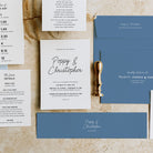 A styled flat-lay of a wedding invitation suite on a textured beige fabric background. The set includes a blue envelope, a white invitation card with black script reading 'Poppy & Christopher', smaller white detail cards with event text, and a gold pocket watch as a decorative prop.
