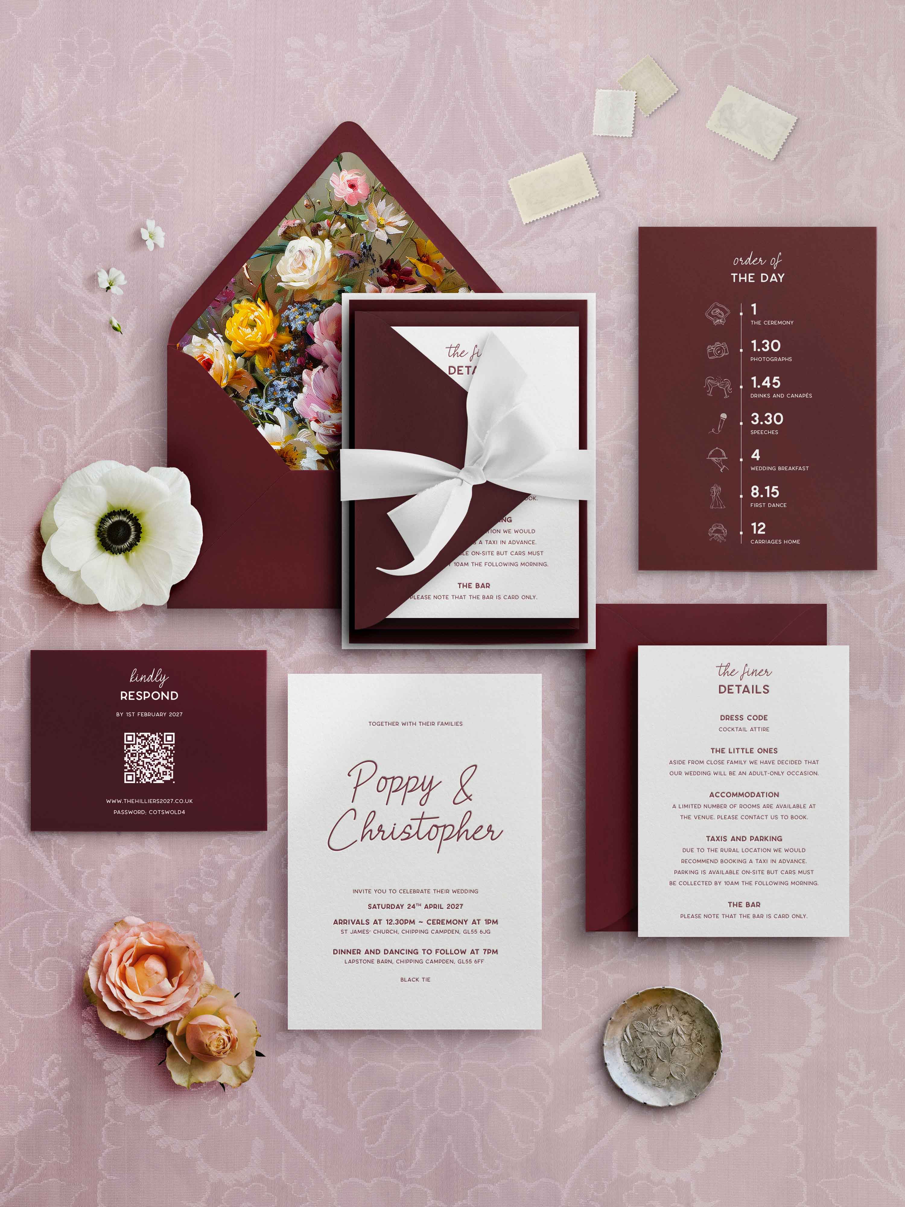 A styled flat lay of a wedding invitation suite on a dusty pink patterned background, including a burgundy letterpress and white invitation card tied with a white ribbon, a floral envelope, a details card, a RSVP card, and decorative flowers.