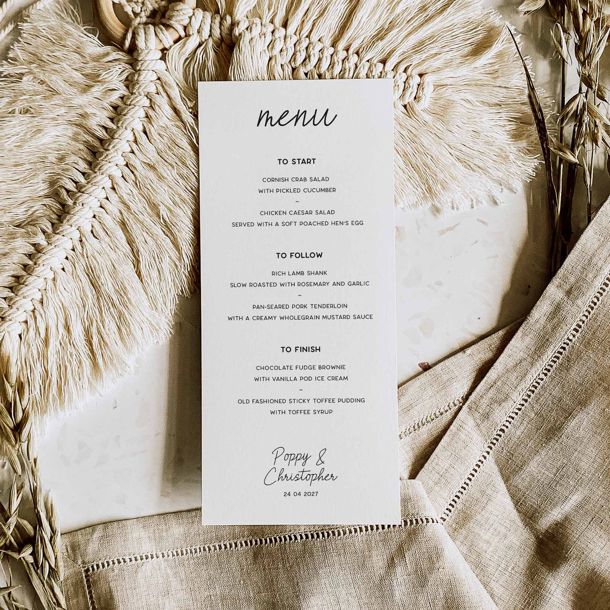 A vertical white paper menu card with black script and serif text, laid flat on a beige linen surface with a cream woven textile and dried botanicals around it. The menu reads 'menu' at the top and includes a to start, to follow, and to finish list with the names 'Poppy & Christopher' and a date at the bottom.
