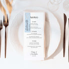 White wedding menu card with a light blue guest name tag and gold paper clip on a white plate next to copper cutlery and glassware