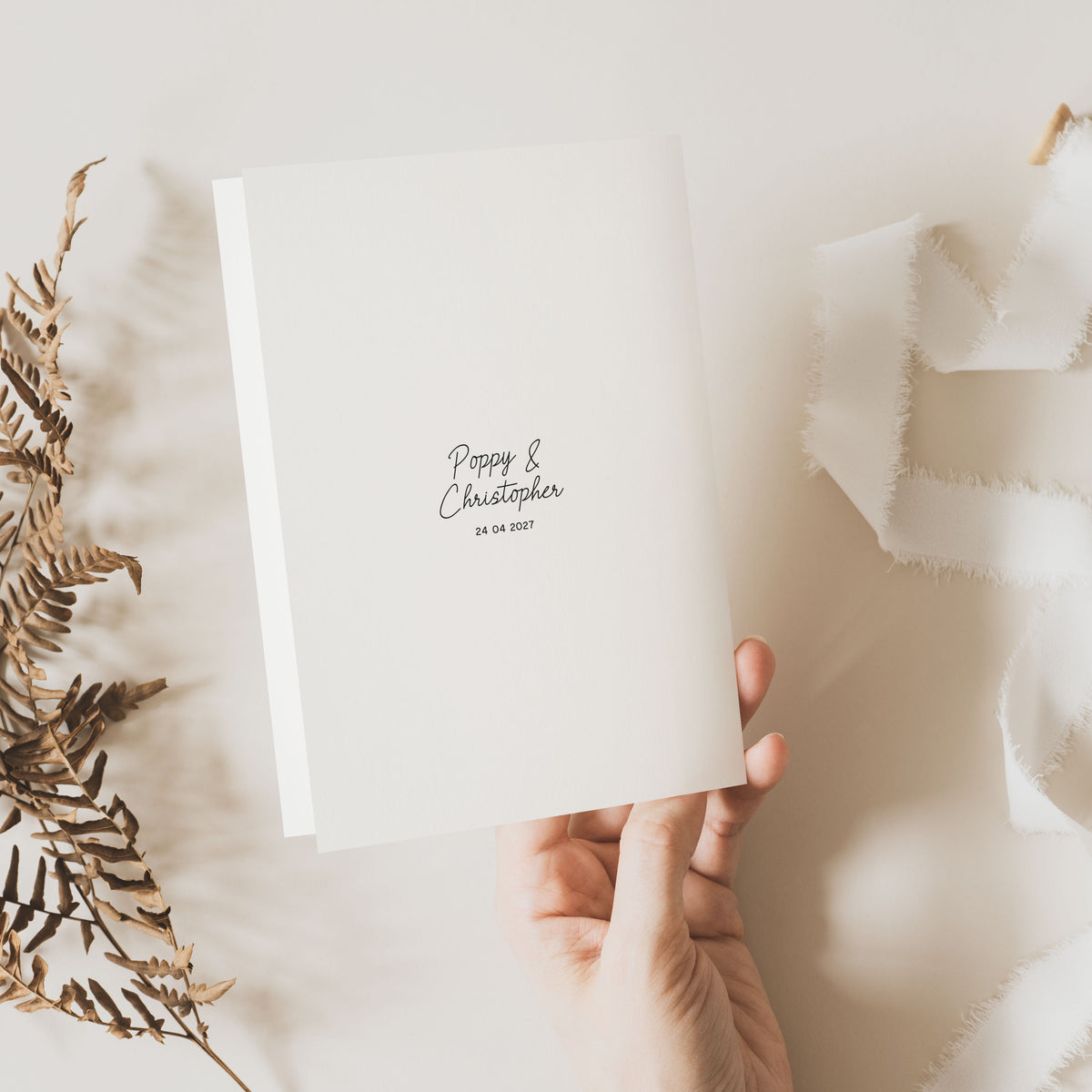 Hand holding a white order of service booklet showing the back cover with a monogram on a light background