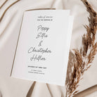 A white wedding order of service booklet with elegant black script text, styled on a draped cream fabric background with dried pampas grass.