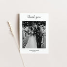 A square white wedding thank-you card with a black-and-white wedding photo in the centre, the word 'thank you' at the top, and personalised text at the bottom, styled on a light beige background with a dried wheat stem.
