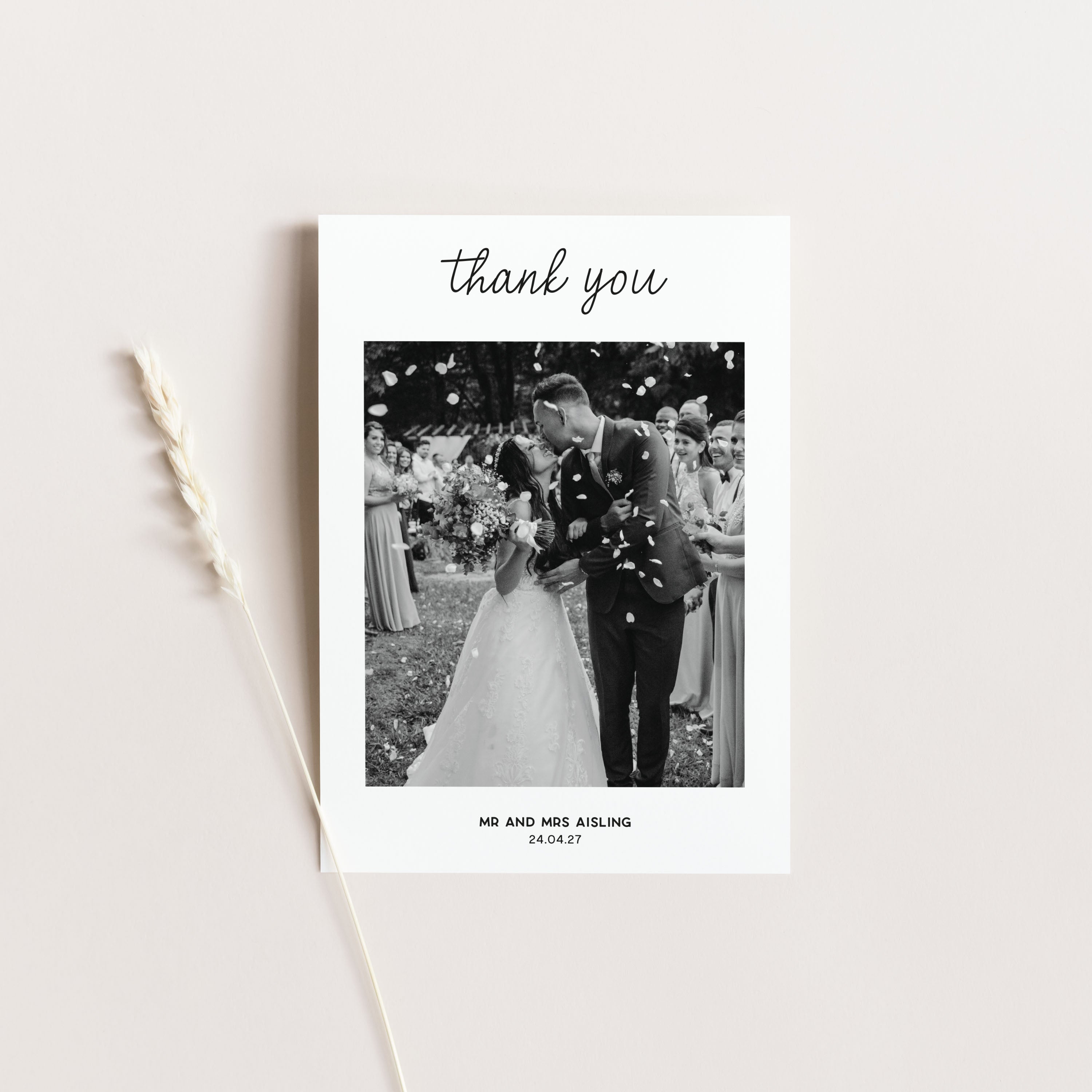 A square white wedding thank-you card with a black-and-white wedding photo in the centre, the word 'thank you' at the top, and personalised text at the bottom, styled on a light beige background with a dried wheat stem.
