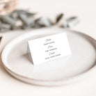 A small white standing place card with black printed text, photographed on a light ceramic plate with a softly blurred neutral background and dried leaves.