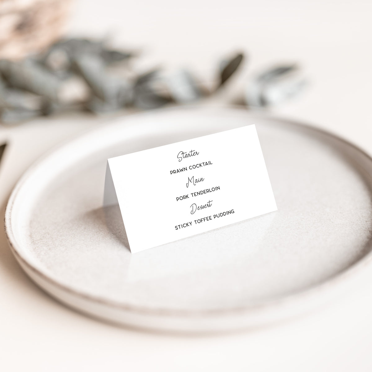 A small white standing place card with black printed text, photographed on a light ceramic plate with a softly blurred neutral background and dried leaves.