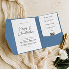 A blue and white wedding invitation card set styled on a textured beige fabric background with a gold wax stamp and white flowers.