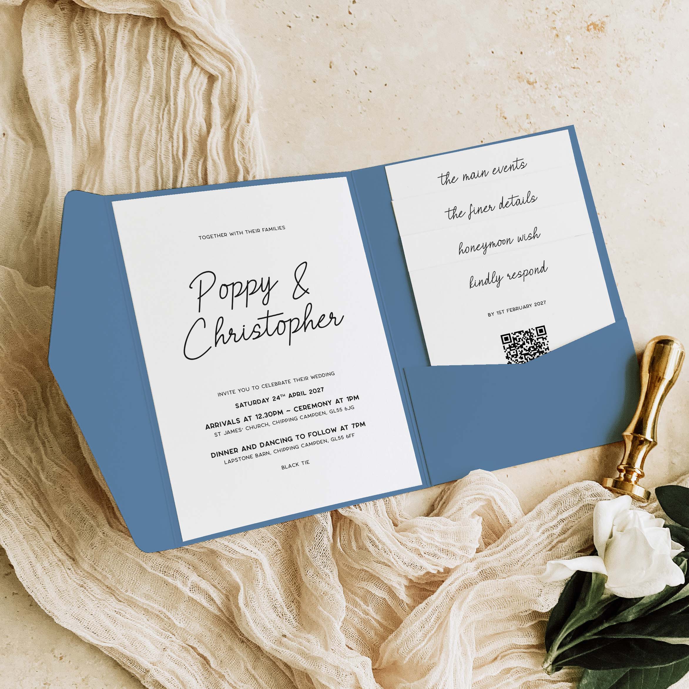 A blue and white wedding invitation card set styled on a textured beige fabric background with a gold wax stamp and white flowers.