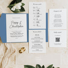 A styled flat-lay of a wedding invitation suite on a light beige marble background with green leaves and white flowers. The set includes a blue envelope, a white invitation card reading 'Poppy & Christopher', an event timeline card, a honeymoon wish card, a dress code card, and a QR code card, along with a gold wax stamp.
