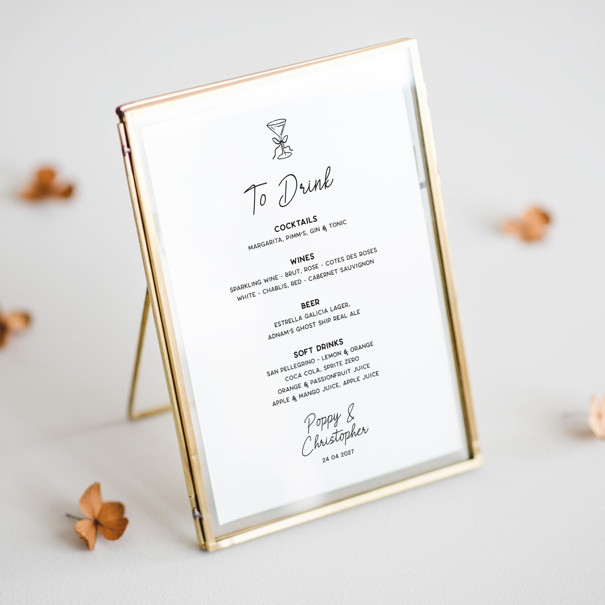 A slim rectangular picture frame with a gold-toned border holds a printed cocktail and wine to-drink menu card, photographed on a light neutral surface with a few small dried flowers around it.