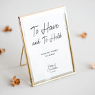 A personalised gold-framed picture frame with a white insert that reads 'To Have and To Hold' and 'Please help yourself to a blanket,' styled on a light gray surface with scattered dried flowers.