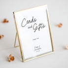 A gold-framed standing sign with a white insert that reads 'Cards and Gifts' and small personalized text below, photographed on a light neutral surface with a few dried flower petals around it.