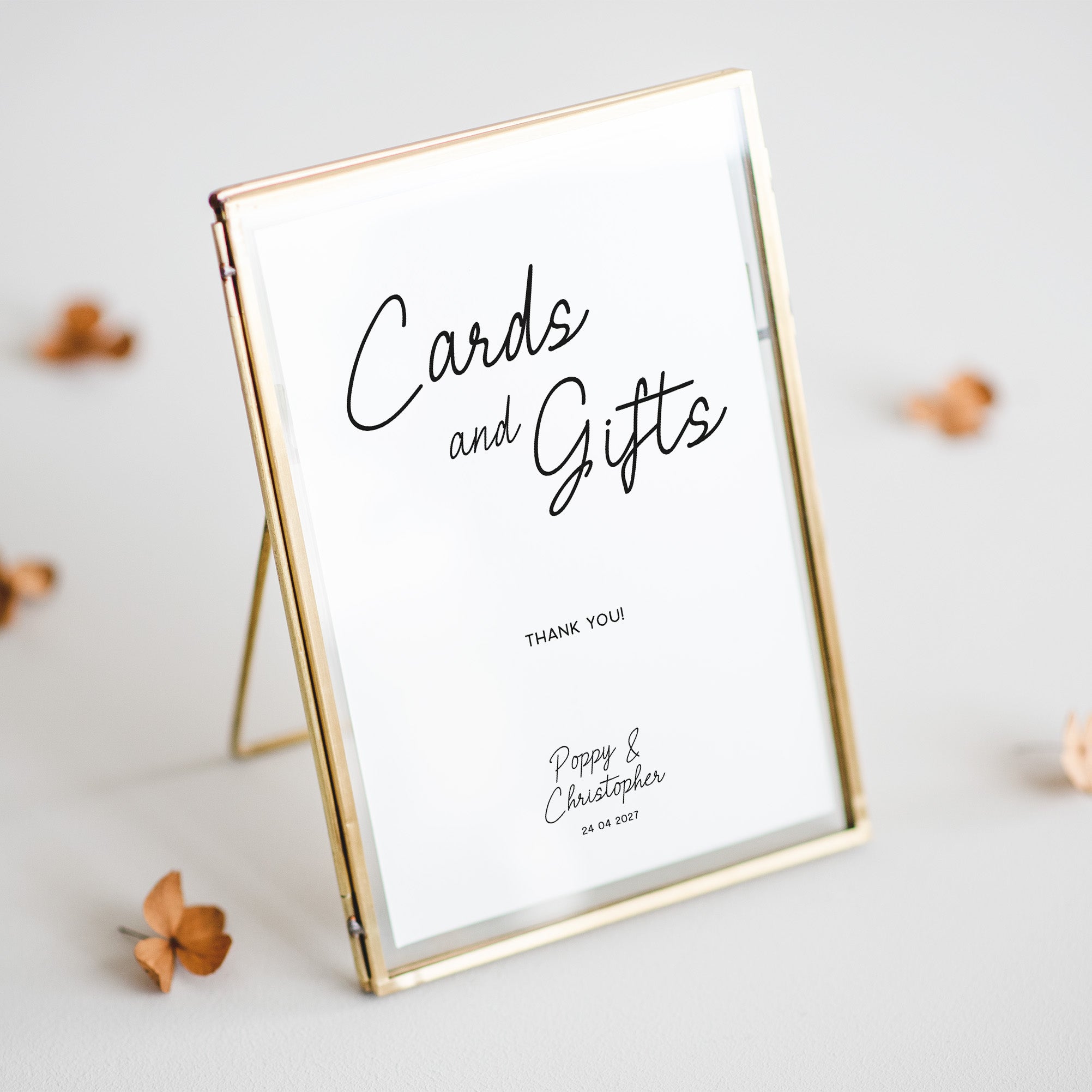 A gold-framed standing sign with a white insert that reads 'Cards and Gifts' and small personalized text below, photographed on a light neutral surface with a few dried flower petals around it.