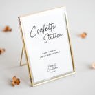 A rectangular gold-framed sign on a small stand with a white insert that reads 'Confetti Station' and smaller text below, photographed on a light neutral background with a few scattered dried flower petals.