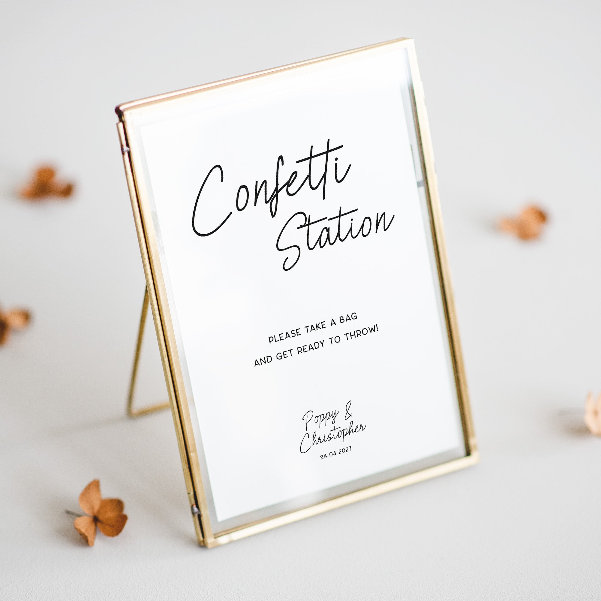A rectangular gold-framed sign on a small stand with a white insert that reads 'Confetti Station' and smaller text below, photographed on a light neutral background with a few scattered dried flower petals.