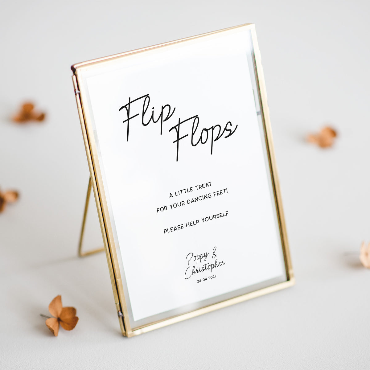A gold-framed standing picture frame with a white insert that reads 'Flip Flops' in black script, photographed on a light neutral surface with a few scattered dried flowers around it.