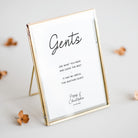 A rectangular picture frame with a thin gold-toned metal border and stand, holding a white printed card that reads 'Gents' with a quote and personalized names/date. The frame is photographed on a light neutral surface with a few small dried flowers around it.