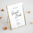 A personalised guest book sign in a gold-framed glass stand, displayed on a light grey surface with scattered dried flowers.