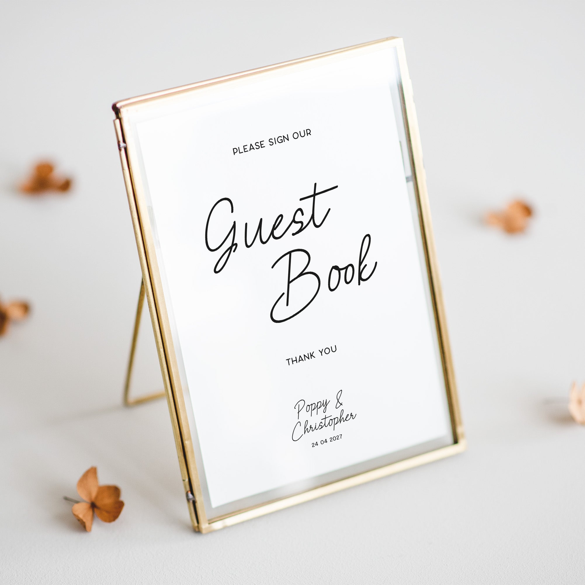 A personalised guest book sign in a gold-framed glass stand, displayed on a light grey surface with scattered dried flowers.