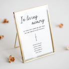 A gold-framed standing picture frame with a white insert printed with the text 'In loving memory' and a small candle illustration, photographed on a light neutral background with a few dried flowers around it.