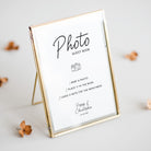 A gold-framed guest book photo frame with a white insert displaying printed instructions and the names 'Pippa & Christopher' with a date, styled on a light gray surface with scattered dried flowers.