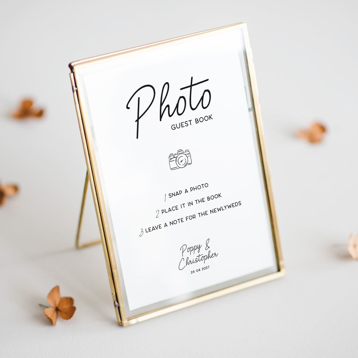 A gold-framed guest book photo frame with a white insert displaying printed instructions and the names 'Pippa & Christopher' with a date, styled on a light gray surface with scattered dried flowers.
