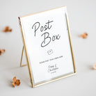 A gold-framed glass post box sign with a white insert that reads 'Post Box' and includes a small envelope icon and personalised text at the bottom, photographed on a light neutral surface with a few dried flowers around it.