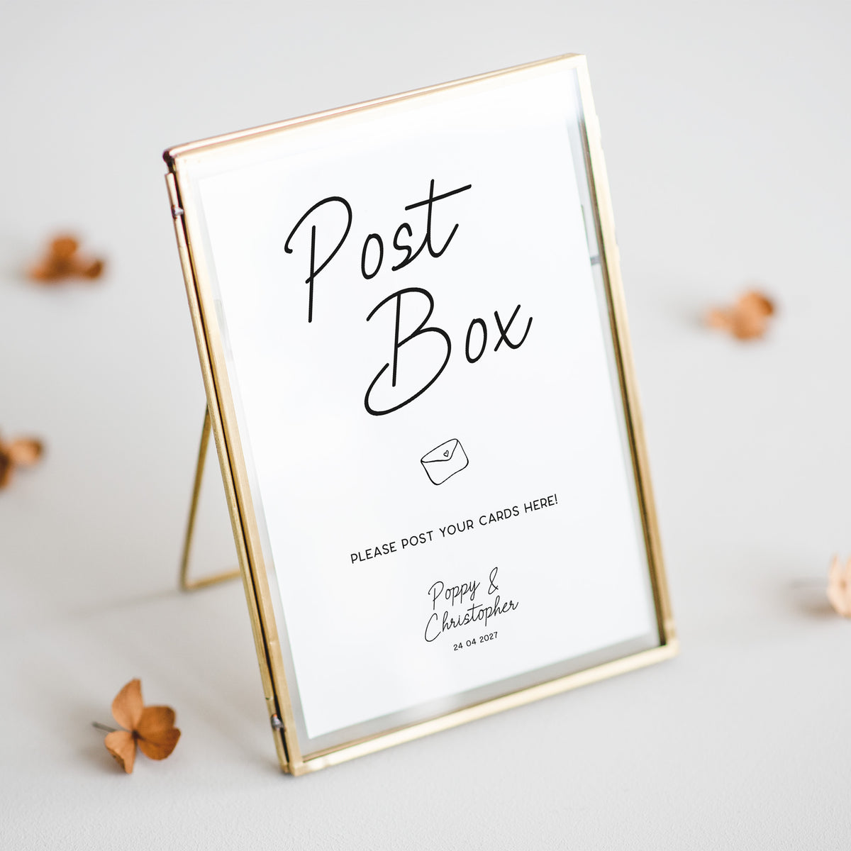 A gold-framed glass post box sign with a white insert that reads 'Post Box' and includes a small envelope icon and personalised text at the bottom, photographed on a light neutral surface with a few dried flowers around it.