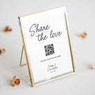 A gold-framed standing photo frame with a white insert that reads 'Share the love' and a QR code, styled on a light surface with scattered dried flowers.