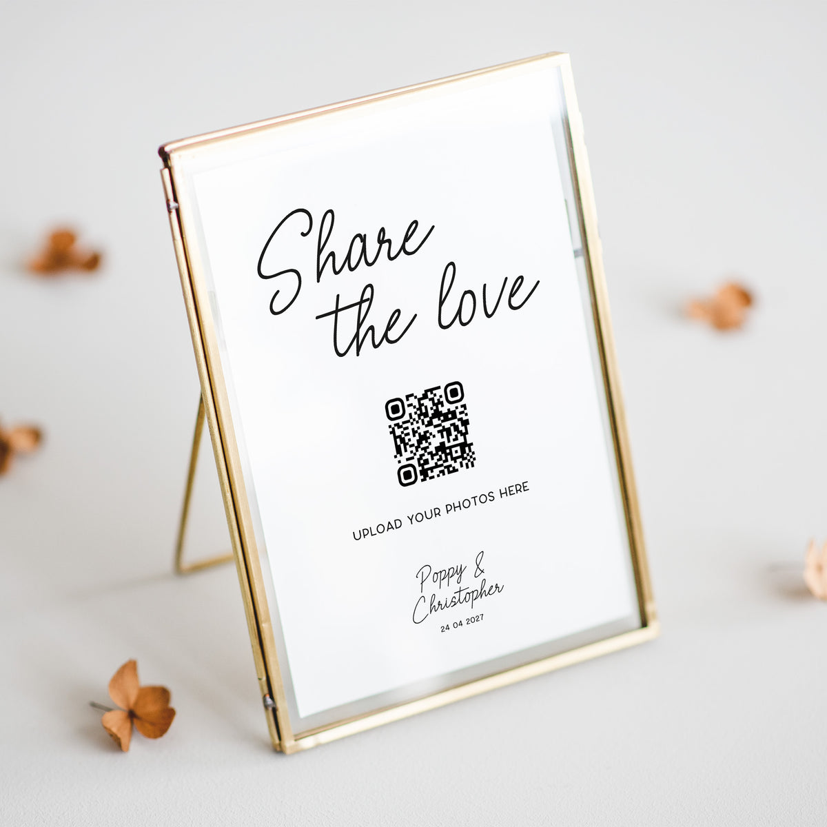 A gold-framed standing photo frame with a white insert that reads 'Share the love' and a QR code, styled on a light surface with scattered dried flowers.