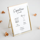 A gold-framed vertical photo frame with a white insert, styled on a light gray surface with scattered dried leaves. The insert features black text reading 'Signature Drinks' with line drawings of a cocktail and a margarita, plus personalized names and date at the bottom.