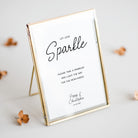 A square mirrored wedding sign with a gold frame and a white insert, standing on a small gold stand. The insert features black script text reading 'Sparkle' and wedding details.
