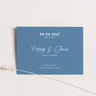 A rectangular blue wedding save-the-date card with white text, photographed on a light neutral background with a thin decorative plant stem crossing the lower left corner.