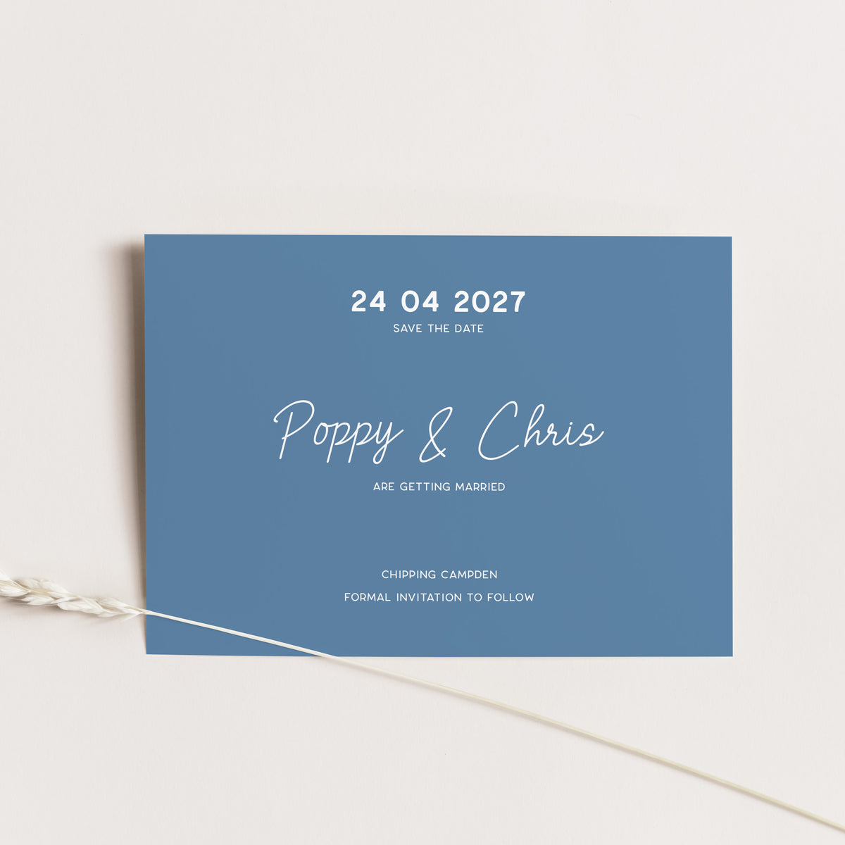 A rectangular blue wedding save-the-date card with white text, photographed on a light neutral background with a thin decorative plant stem crossing the lower left corner.