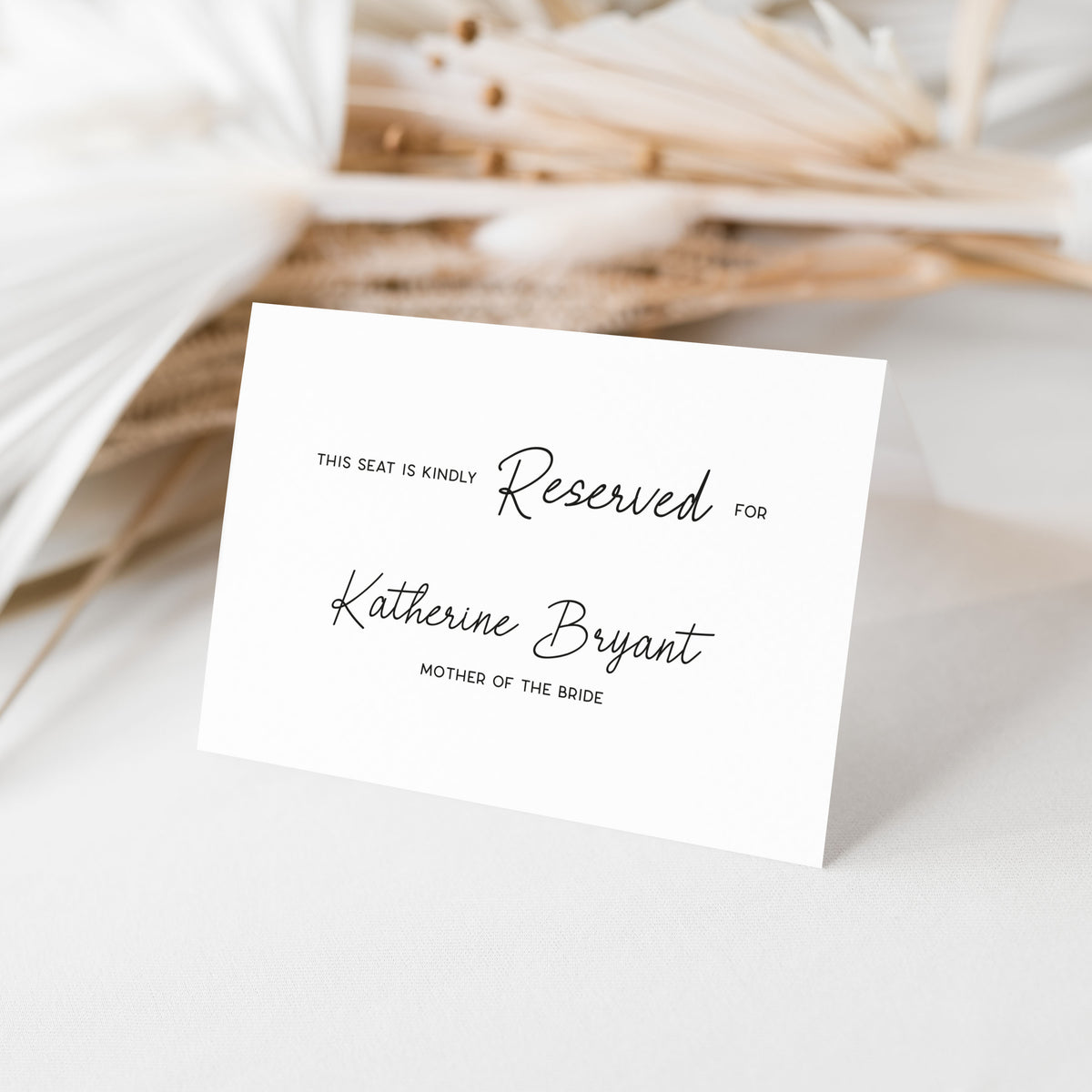 A white reserved seat card with black script text reading 'Reserved for Katherine Bryant, mother of the bride,' photographed standing on a light surface with soft dried floral props in the background.