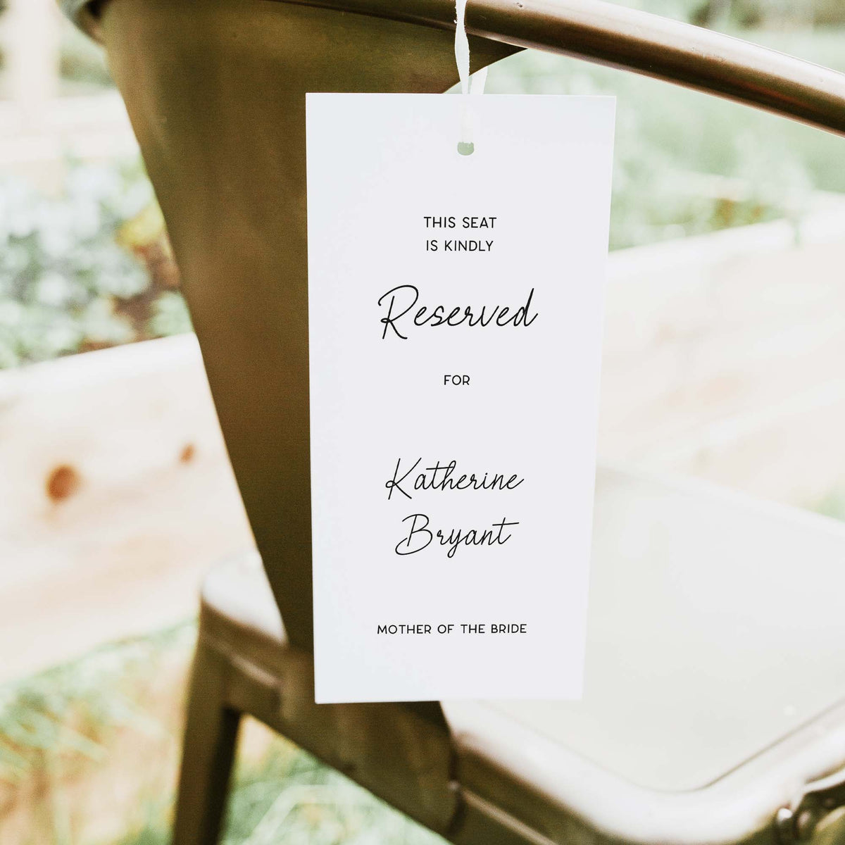A white reserved seat tag hanging from a metal chair in an outdoor setting. The tag features elegant black script and serif text reading 'This seat is kindly Reserved for Katherine Bryant, Mother of the Bride'.