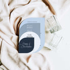 A flat-lay photograph of a custom wedding invitation card with a modern geometric design in light blue, gray, white, and navy. The card is styled on draped beige fabric next to two clear ribbed glass tumblers.