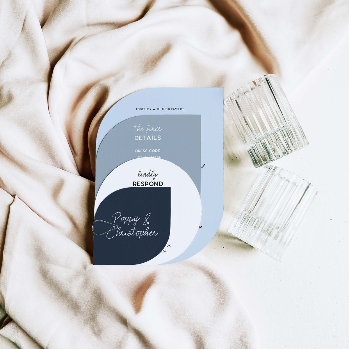 A flat-lay photograph of a custom wedding invitation card with a modern geometric design in light blue, gray, white, and navy. The card is styled on draped beige fabric next to two clear ribbed glass tumblers.