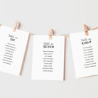 Three vertical white cards with printed table numbers hang from a string between two wooden clothespins against a light gray background.