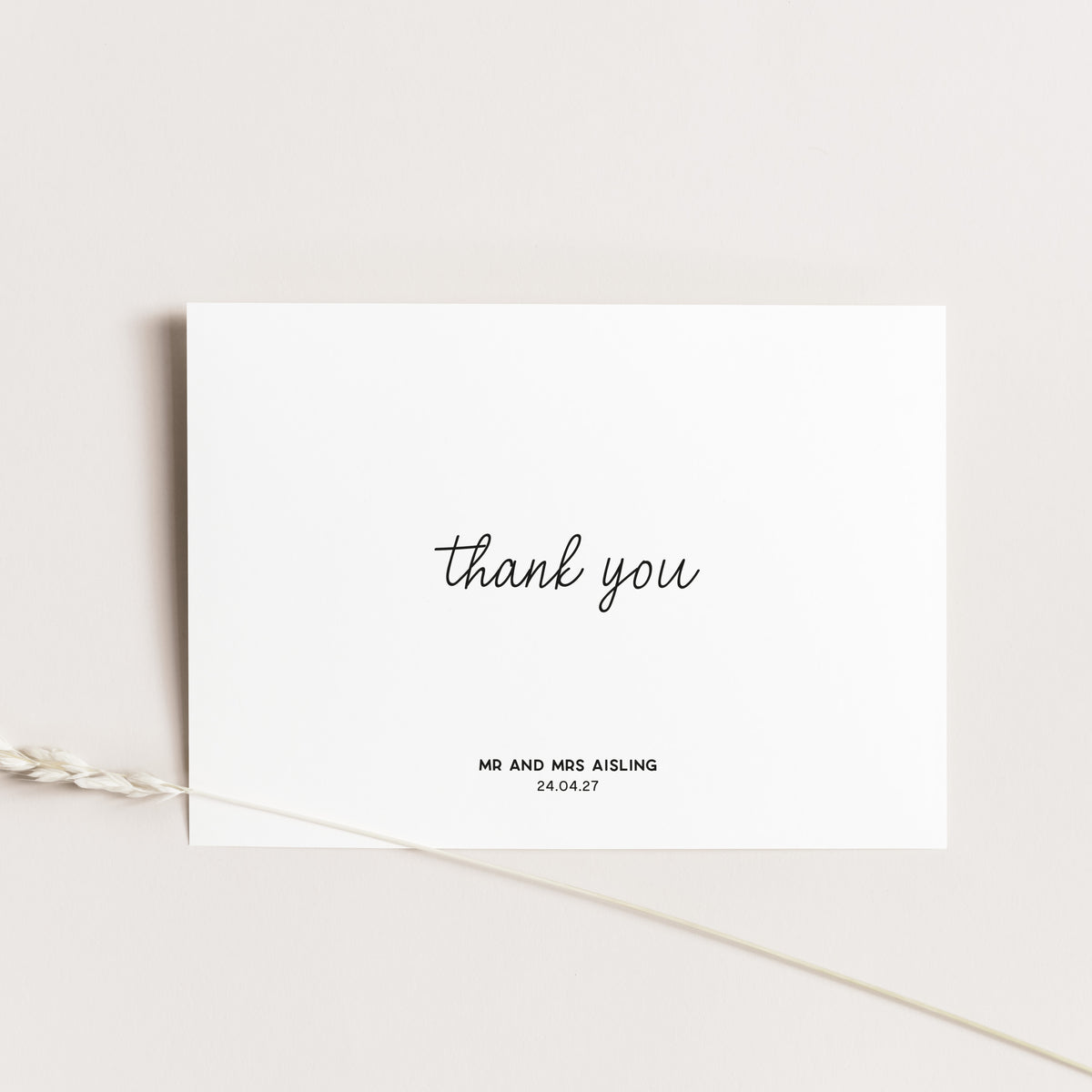 A minimalist white thank-you card with black script text reading 'thank you' and smaller personalized text below, photographed on a light neutral background with a dried plant stem crossing the lower left corner.