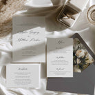 A styled flat-lay of a wedding invitation suite on white satin fabric, including an invitation card, a menu card, and a website card, alongside a gray envelope, a silver ring box with a ring, and dried flowers.